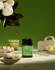A studio photoshoot featuring a jar of premium New Zealand BeeZ Kanuka Honey on a table with a linen tablecloth, slices of sourdough bread and a ceramic plate of New Zealand butter. A vase of magnolias are in the background.