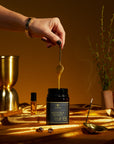 A studio photoshoot of a jar of BeeNZ Premium New Zealand Manuka Honey with a woman's hand holding a teaspoon full of honey dripping into the jar. Incense, an oil roller and a vase of manuka are in the frame along with a metallic gold tablecloth.