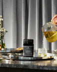 A jar of Premium New Zealand Manuka Honey made by BeeNZ with a woman's hand pouring a herbal tea from a glass teapot into a glass teacup