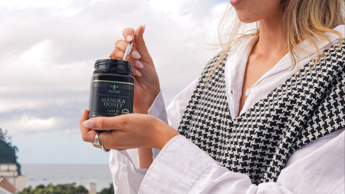 A woman outdoors holding a small jar, wearing a white blouse and a black-and-white houndstooth vest, with a cloudy sky and trees in the background.