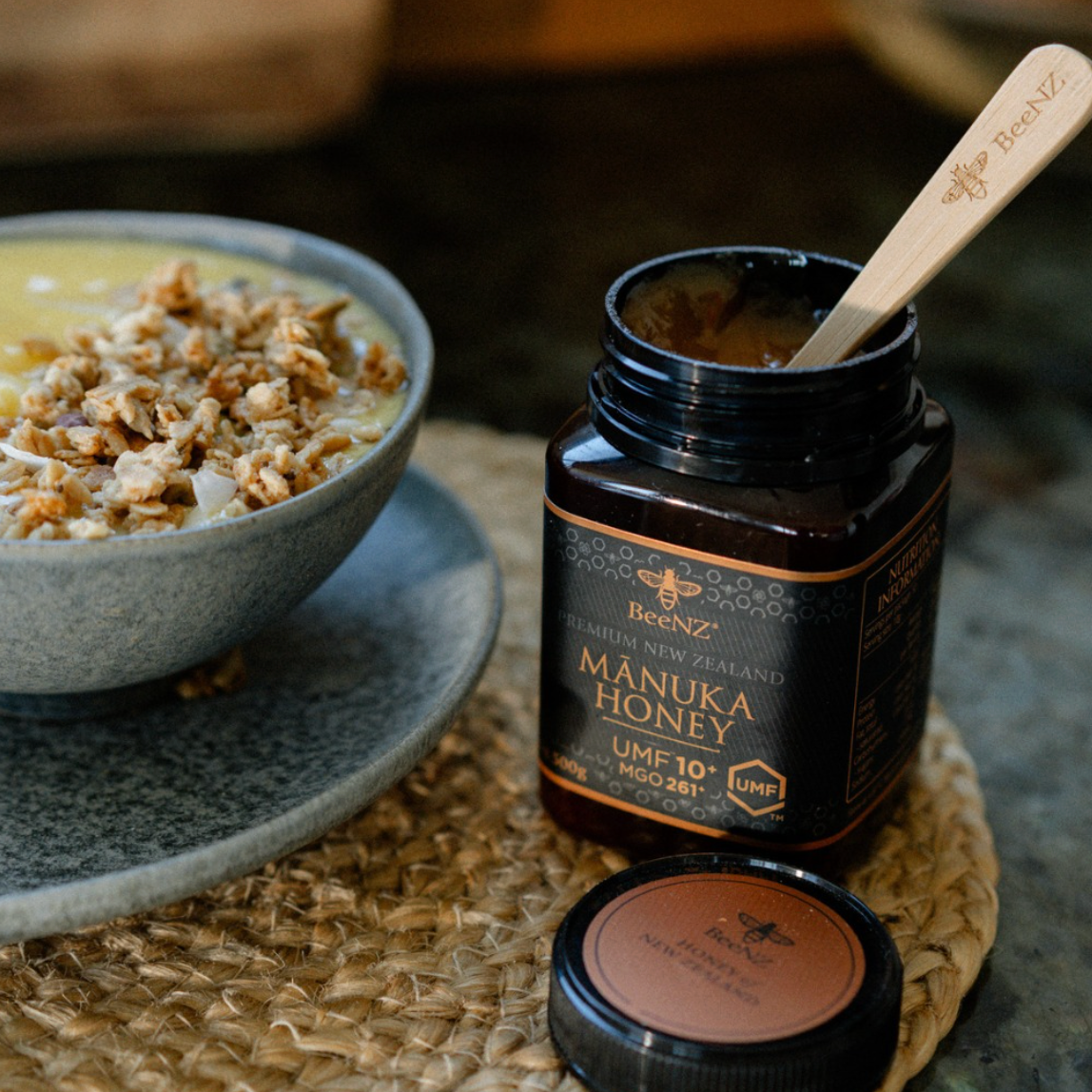 Manuka honey umf10+ jar with a spoon on a woven mat next to a bowl of cereal.