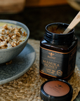 Manuka honey umf10+ jar with a spoon on a woven mat next to a bowl of cereal.